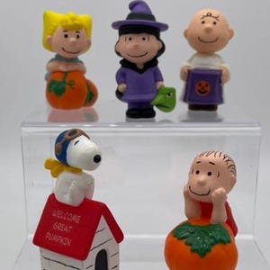 May include: A set of five Peanuts character figurines including Sally sitting on a pumpkin, Lucy in a purple witch costume, Charlie Brown holding a purple trick-or-treat bag, Snoopy on a red doghouse with the text WELCOME GREAT PUMPKIN, and Linus.