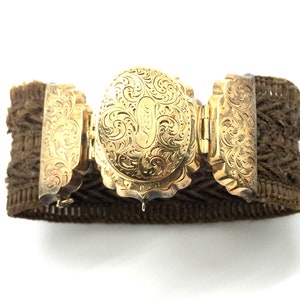 May include: A gold-toned bracelet with an ornate, engraved clasp. The bracelet is made of a brown, woven material.