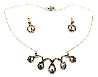 Puede incluir: Un conjunto de collar y pendientes de oro con una cadena delicada y un colgante con múltiples piedras de granate rojo. El colgante tiene la forma de una serie de círculos conectados.