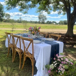 May include: A white tablecloth with a blue runner is set on a wooden table with six chairs. A bouquet of pink and yellow flowers is in a white vase in the centre of the table. The table is set with plates, cutlery, and glasses. The table is outdoors under a tree with a green grassy field in the background.