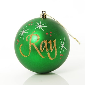 Op de afbeelding: Groene kerstbal met de naam "Ray" geschreven in gouden glitter. De kerstbal is versierd met witte glittersterren en rode glitterpunten.
