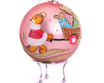 Op de afbeelding: Roze glazen kerstbal met een geschilderd ontwerp van een teddybeer die een kar vol cadeautjes trekt. De bal staat op een zilveren standaard.
