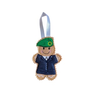 May include: A gingerbread man ornament with a green beret and a navy blue suit. The ornament has a light brown body, a white ribbon for hanging, and a smiling face. The beret has a yellow and green emblem.