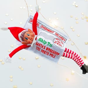 May include: A festive Christmas elf doll with a red hat, arms, and legs, and a smiling face. The elf is holding a grey envelope that reads "Special Overnight Delivery" and "Ship To: Santa's Workshop The North Pole."