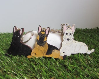 May include: Three wooden dog keychain charms. The charms are of German Shepherd dogs. One is black, one is brown and black, and one is white. The charms are on a green grassy surface.