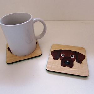 May include: Two wooden coasters with a green border. One coaster features a brown dog face with black eyes and nose. The other coaster is empty.