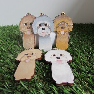 May include: Five wooden keychains shaped like poodles in different colors. The keychains are brown, gray, white, and two shades of tan. The poodles have painted faces with black eyes and pink noses.