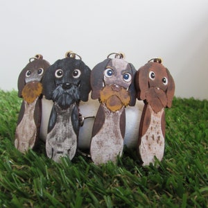 Puede incluir: Cuatro llaveros de madera con forma de perro pintados en diferentes colores y patrones. Los perros están todos de pie sobre una superficie verde.