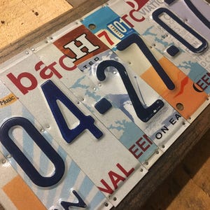 K&ouml;nnte beinhalten: Ein Wandkunstwerk mit Holzrahmen aus wiederverwendeten Nummernschildern. Die Nummernschilder sind auf dem Holz vernietet und zeigen die Zahlen "04-27-0". Die Nummernschilder sind in verschiedenen Farben und Designs gehalten.