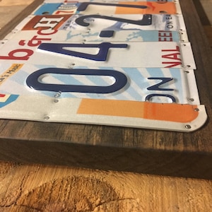 K&ouml;nnte beinhalten: Ein rechteckiges Holzschild mit einer Collage aus alten Nummernschildern. Die Schilder sind auf dem Holz vernietet und zeigen eine Vielzahl von Farben, Zahlen und Buchstaben. Das Schild hat einen dunkelbraunen Holzrahmen.