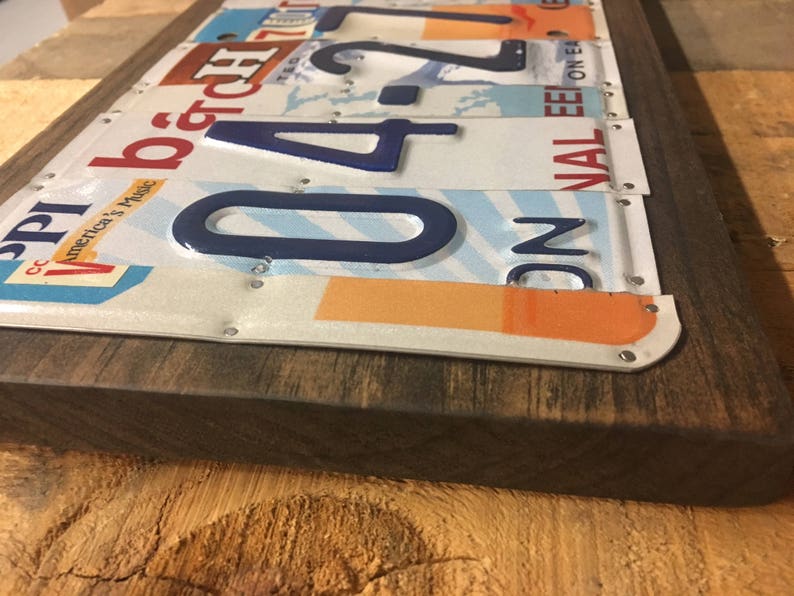 K&ouml;nnte beinhalten: Ein rechteckiges Holzschild mit einer Collage aus alten Nummernschildern. Die Schilder sind auf dem Holz vernietet und zeigen eine Vielzahl von Farben, Zahlen und Buchstaben. Das Schild hat einen dunkelbraunen Holzrahmen.
