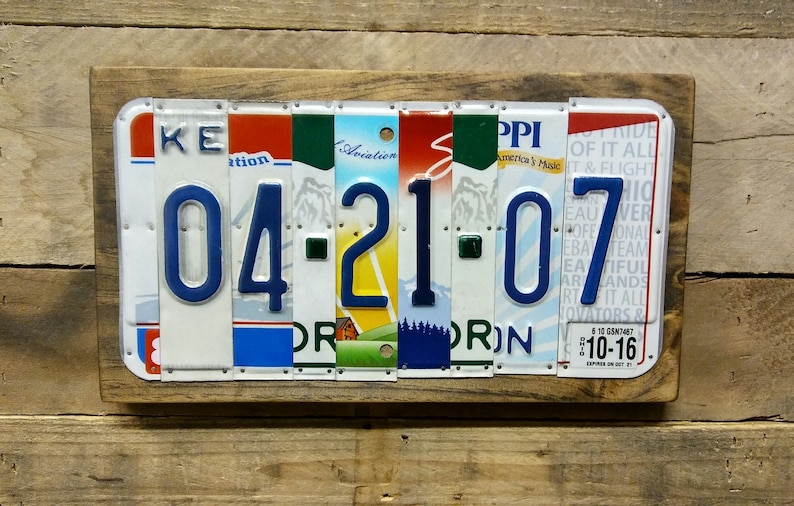 K&ouml;nnte beinhalten: Ein personalisiertes Wandbild mit dem Datum "04-21-07", das aus wiederverwerteten Nummernschildern gefertigt wurde. Das Datum wird in blauen Ziffern vor einem Hintergrund aus farbigen Nummernschildfragmenten dargestellt, eingerahmt in einem rustikalen Holzrahmen.