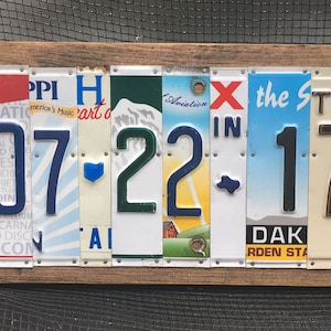 K&ouml;nnte beinhalten: Ein Wandkunstwerk mit Holzrahmen, das das Datum "07-22-17" unter Verwendung von Nummernschildern anzeigt. Die Zahlen sind in verschiedenen Farben und Stilen gehalten, wobei der Hintergrund der Schilder verschiedene Bundesstaaten und Designs zeigt.