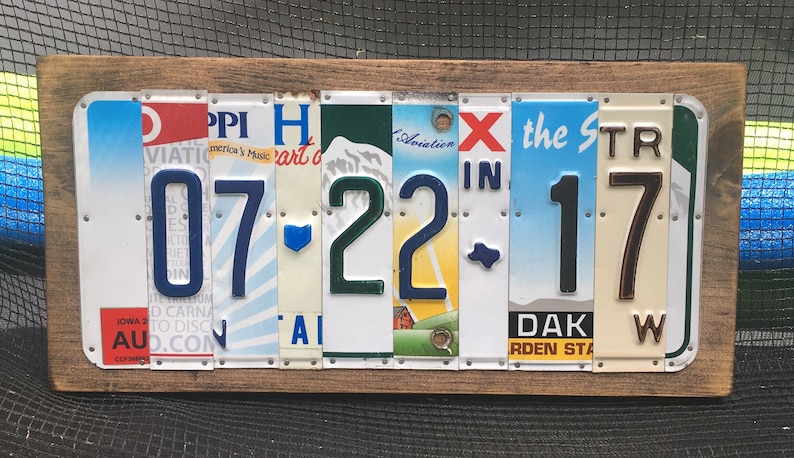 K&ouml;nnte beinhalten: Ein Wandkunstwerk mit Holzrahmen, das das Datum "07-22-17" unter Verwendung von Nummernschildern anzeigt. Die Zahlen sind in verschiedenen Farben und Stilen gehalten, wobei der Hintergrund der Schilder verschiedene Bundesstaaten und Designs zeigt.