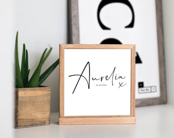 Puede incluir: Una impresión enmarcada en blanco con el nombre Aurelia escrito en cursiva negra y la fecha 07.09.2021 debajo. La impresión está sobre una repisa de madera.