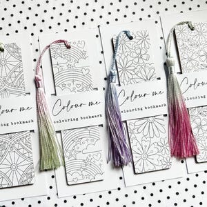 Könnte beinhalten: Vier weiße Mal-Lesezeichen mit bunten Quasten. Jedes Lesezeichen zeigt ein anderes Schwarz-Weiß-Design, darunter geometrische Muster, Blumen, Regenbogen und Sterne. Der Text "Colour me colouring bookmark" ist auf jedem Lesezeichen aufgedruckt.