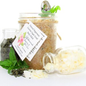 An 8 oz jar of peppermint and coconut cane sugar scrub offers a refreshing and hydrating experience for your skin. The sugar scrub is centered, near bright green sprigs of peppermint, dried mint and coconut flakes are scattered in the arrangement.
