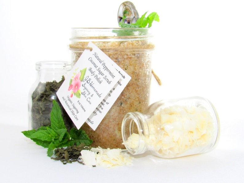 An 8 oz jar of peppermint and coconut cane sugar scrub offers a refreshing and hydrating experience for your skin. The sugar scrub is centered, near bright green sprigs of peppermint, dried mint and coconut flakes are scattered in the arrangement.