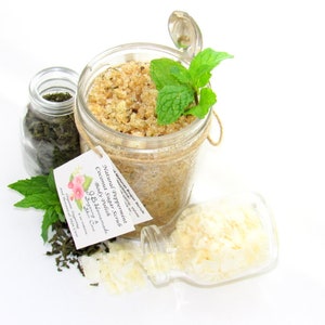An 8 oz jar of peppermint and coconut cane sugar scrub offers a refreshing and hydrating experience for your skin. The sugar scrub is centered, near bright green sprigs of peppermint, dried mint and coconut flakes are scattered in the arrangement.