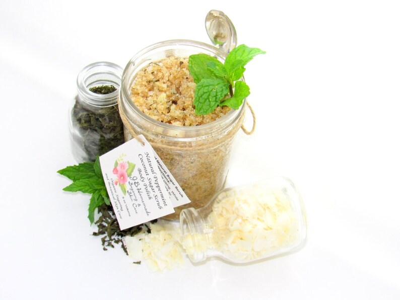 An 8 oz jar of peppermint and coconut cane sugar scrub offers a refreshing and hydrating experience for your skin. The sugar scrub is centered, near bright green sprigs of peppermint, dried mint and coconut flakes are scattered in the arrangement.