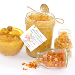 An 8 oz jar of citrus sugar scrub, with orange & calendula, for a radiant glow. The jar is centered by 2 small jars, 1 with orange zest, 1 with calendula spilling on a white table. A bowl of sugar scrub, with calendula petals and an orange slice.