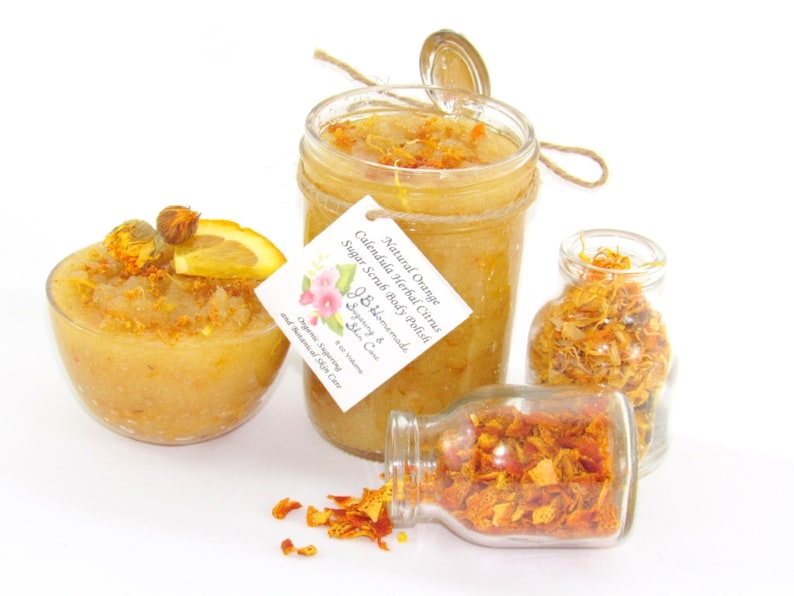 An 8 oz jar of citrus sugar scrub, with orange & calendula, for a radiant glow. The jar is centered by 2 small jars, 1 with orange zest, 1 with calendula spilling on a white table. A bowl of sugar scrub, with calendula petals and an orange slice.