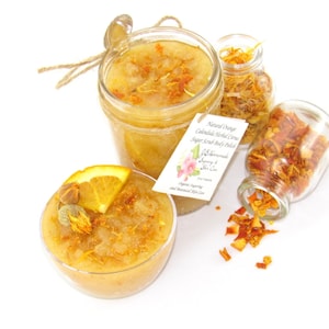 An 8 oz jar of citrus sugar scrub, with orange & calendula, for a radiant glow. The jar is centered by 2 small jars, 1 with orange zest, 1 with calendula spilling on a white table. A bowl of sugar scrub, with calendula petals and an orange slice.