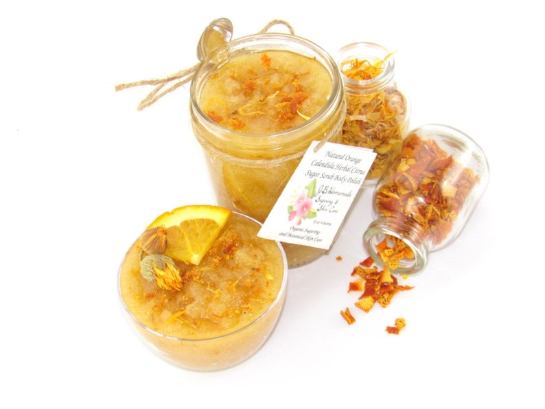 An 8 oz jar of citrus sugar scrub, with orange & calendula, for a radiant glow. The jar is centered by 2 small jars, 1 with orange zest, 1 with calendula spilling on a white table. A bowl of sugar scrub, with calendula petals and an orange slice.