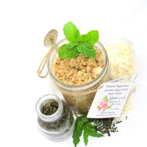 An 8 oz jar of peppermint and coconut cane sugar scrub offers a refreshing and hydrating experience for your skin. The sugar scrub is centered, near bright green sprigs of peppermint, dried mint and coconut flakes are scattered in the arrangement.
