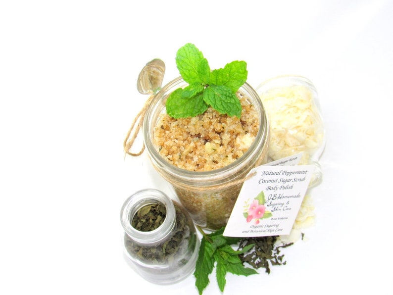 An 8 oz jar of peppermint and coconut cane sugar scrub offers a refreshing and hydrating experience for your skin. The sugar scrub is centered, near bright green sprigs of peppermint, dried mint and coconut flakes are scattered in the arrangement.