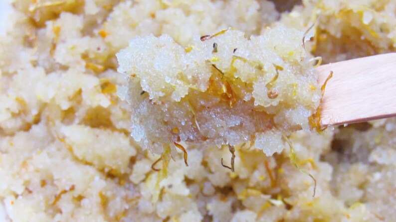 Close-up view of orange calendula sugar scrub in a bowl with a wooden spatula, highlighting its grainy texture and orange color.