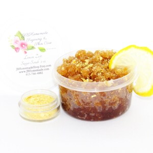 A 2 oz tub of energizing lemon zest raw and cane sugar scrub, zingy and refreshing, leaves your skin feeling invigorated. The scrub is centered, topped with lemon zest and a slice, with a bowl filled to the brim with dried lemon zest to the left.