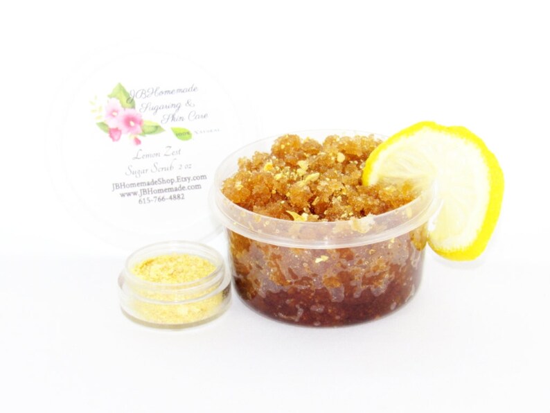 A 2 oz tub of energizing lemon zest raw and cane sugar scrub, zingy and refreshing, leaves your skin feeling invigorated. The scrub is centered, topped with lemon zest and a slice, with a bowl filled to the brim with dried lemon zest to the left.