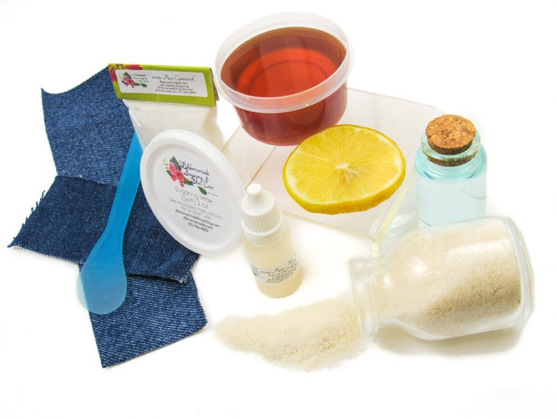 A 2 oz tub of JBHomemade Sugaring Wax with its included cornstarch, aloe vera, denim strips & applicator next to a slice of lemon, a glass jar filled with clear water, and a jar tipped over, spilling cane sugar, accentuating the natural ingredients.