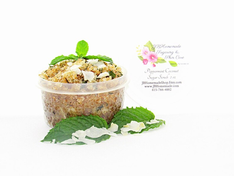 A 2 oz tub of cooling peppermint and coconut cane sugar scrub offers a refreshing and hydrating experience for your skin. The sugar scrub is centered, near bright green sprigs of peppermint and coconut flakes are scattered throughout the arrangement.