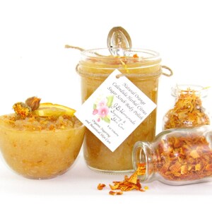 An 8 oz jar of citrus sugar scrub, with orange & calendula, for a radiant glow. The jar is centered by 2 small jars, 1 with orange zest, 1 with calendula spilling on a white table. A bowl of sugar scrub, with calendula petals and an orange slice.