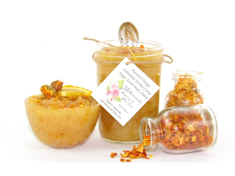 An 8 oz jar of citrus sugar scrub, with orange & calendula, for a radiant glow. The jar is centered by 2 small jars, 1 with orange zest, 1 with calendula spilling on a white table. A bowl of sugar scrub, with calendula petals and an orange slice.