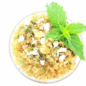 A glass bowl of JBHomemade Sugaring and Skin Care's Natural Peppermint Coconut Sugar Body Scrub with sprinkles of coconut shavings, dried mint and a fresh mint sprig on top.