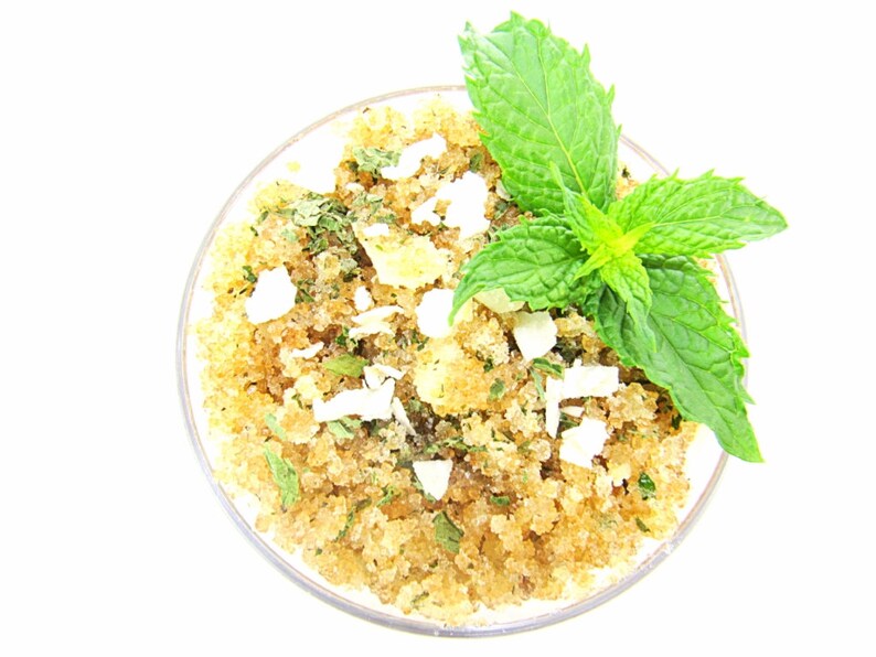 A glass bowl of JBHomemade Sugaring and Skin Care's Natural Peppermint Coconut Sugar Body Scrub with sprinkles of coconut shavings, dried mint and a fresh mint sprig on top.