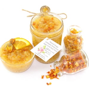 An 8 oz jar of citrus sugar scrub, with orange & calendula, for a radiant glow. The jar is centered by 2 small jars, 1 with orange zest, 1 with calendula spilling on a white table. A bowl of sugar scrub, with calendula petals and an orange slice.