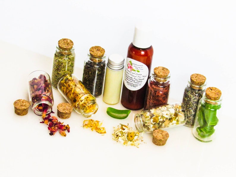An all-natural facial cleanser in an amber bottle surrounded by eight small corked glass bottles containing sprinkles of Calendula, Rose, Lavender, Chamomile, Rosemary, Aloe Vera, Sandalwood and Patchouli ingredients and sprinkles of the same.