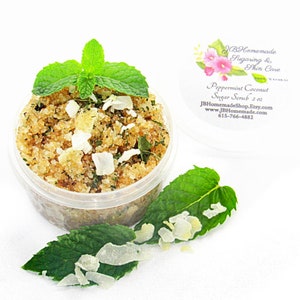 A 2 oz tub of cooling peppermint and coconut cane sugar scrub offers a refreshing and hydrating experience for your skin. The sugar scrub is centered, near bright green sprigs of peppermint and coconut flakes are scattered throughout the arrangement.