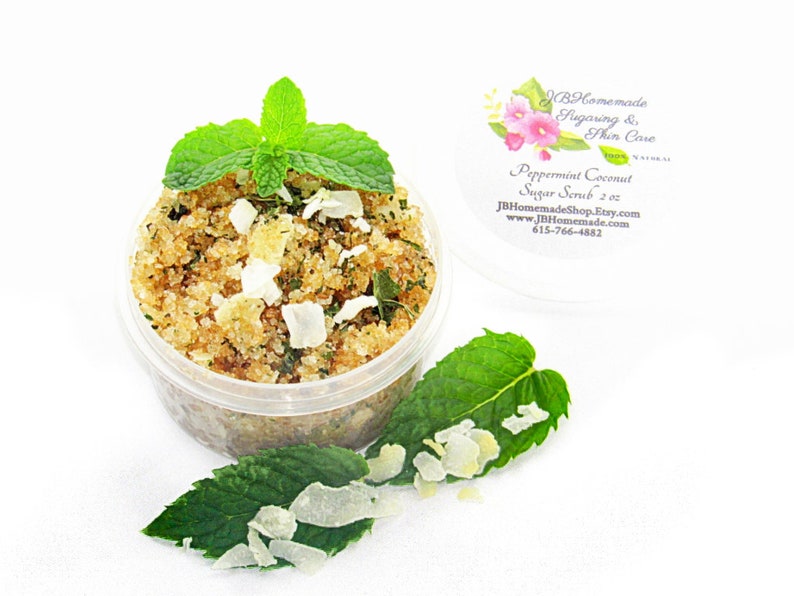 A 2 oz tub of cooling peppermint and coconut cane sugar scrub offers a refreshing and hydrating experience for your skin. The sugar scrub is centered, near bright green sprigs of peppermint and coconut flakes are scattered throughout the arrangement.