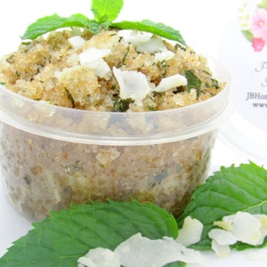 A 2 oz tub of cooling peppermint and coconut cane sugar scrub offers a refreshing and hydrating experience for your skin. The sugar scrub is centered, near bright green sprigs of peppermint and coconut flakes are scattered throughout the arrangement.