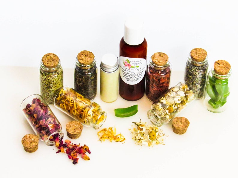 An all-natural facial cleanser in an amber bottle surrounded by eight small corked glass bottles containing sprinkles of Calendula, Rose, Lavender, Chamomile, Rosemary, Aloe Vera, Sandalwood and Patchouli ingredients and sprinkles of the same.