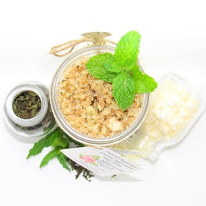 An 8 oz jar of peppermint and coconut cane sugar scrub offers a refreshing and hydrating experience for your skin. The sugar scrub is centered, near bright green sprigs of peppermint, dried mint and coconut flakes are scattered in the arrangement.