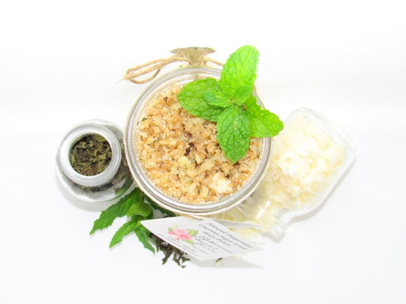 An 8 oz jar of peppermint and coconut cane sugar scrub offers a refreshing and hydrating experience for your skin. The sugar scrub is centered, near bright green sprigs of peppermint, dried mint and coconut flakes are scattered in the arrangement.