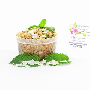 A 2 oz tub of cooling peppermint and coconut cane sugar scrub offers a refreshing and hydrating experience for your skin. The sugar scrub is centered, near bright green sprigs of peppermint and coconut flakes are scattered throughout the arrangement.