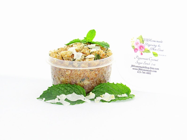 A 2 oz tub of cooling peppermint and coconut cane sugar scrub offers a refreshing and hydrating experience for your skin. The sugar scrub is centered, near bright green sprigs of peppermint and coconut flakes are scattered throughout the arrangement.