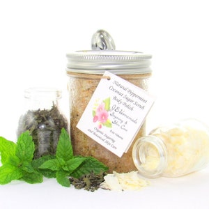 An 8 oz jar of peppermint and coconut cane sugar scrub offers a refreshing and hydrating experience for your skin. The sugar scrub is centered, near bright green sprigs of peppermint, dried mint and coconut flakes are scattered in the arrangement.